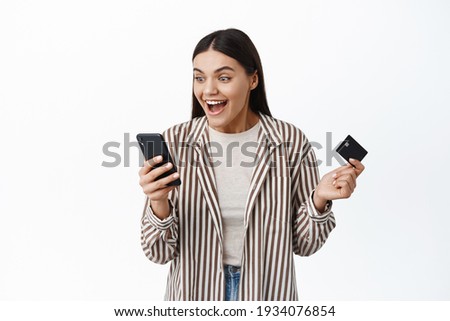 Excited and surprised woman winning money cashback, holding plastic credit card and staring happy at smartphone, standing over white background