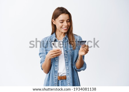 Online shopping. Smiling good-looking woman paying for order, using plastic credit card to pay with mobile phone, standing against white background