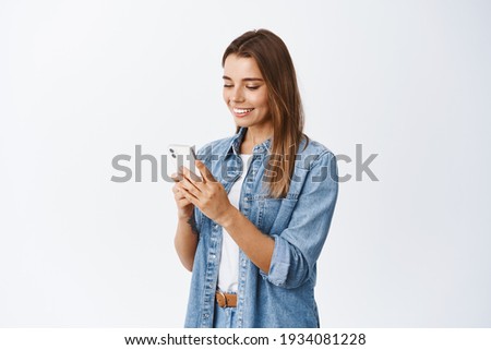 Portrait of young blond woman chatting on social media, holding smartphone and texting with pleased smile, standing against white background