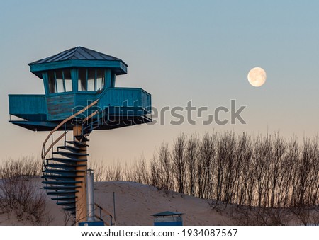 full moon in the morning and a tower for beach rescuers in blue with stairs and beach sand bushes