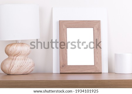 A white photo frame on a wooden table, a standing wood lamp and a white candle next to it