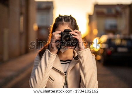 Young girl with black hair, Caucasian, dressed in beige windbreaker, black and white striped t-shirt, taking a picture with her reflex camera in a sunset in the street using ffp2 mask