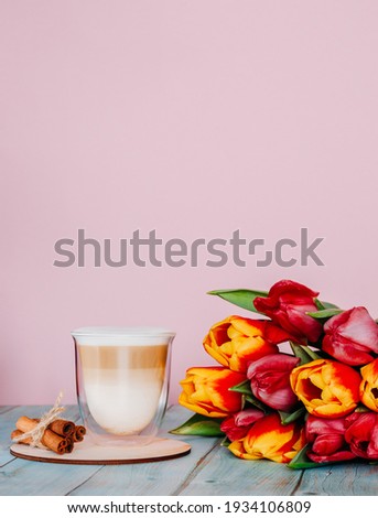 Cappuccino coffee in a double glass on a pink background and a wooden stand with cinnamon and a bouquet of bright beautiful tulips.