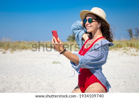 Happy young woman in red bikini using smartphone for video call, standing on beach outdoor, wearing summer straw hat and sunglasses.Making selfie, sharing data on social media.Travel vacation concept