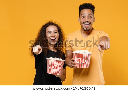 Young couple two friends together family african smiling man woman in black t-shirt hold popcorn snack takeaway bucket point index finger camera on you isolated on yellow background studio portrait.