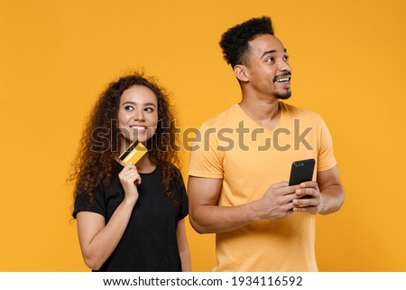Young couple two friends together family african dreamful man woman 20s in black t-shirt holding mobile phone credit bank card shop online look aside isolated on yellow background studio portrait