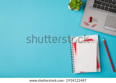 Top view photo of workplace with laptop red chancellery notebooks plant on isolated blue background with blank space