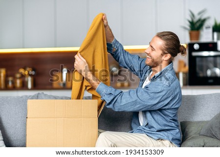 Smiling attractive man unpacked his parcel, happy about getting a long expected order. Caucasian modern guy shopping in internet stores, buying new clothes online, online shopping concept Royalty-Free Stock Photo #1934153309
