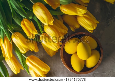 Easter eggs in wooden bowl and bouquet of bright yellow tulips on gray concrete surface. Flat lay, top view