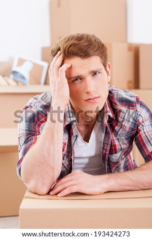 Tired of moving. Depressed young man sitting on the floor and holding hand in hair while cardboard boxes laying on the background