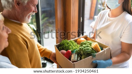 Food delivery or donation concept. Young woman in gloves and protective face mask delivers a box of food elderly couple in home Royalty-Free Stock Photo #1934250461