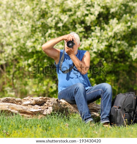 old photographer sitting on a fallen tree and photographs. Elderly man enjoys traveling and photography nature.