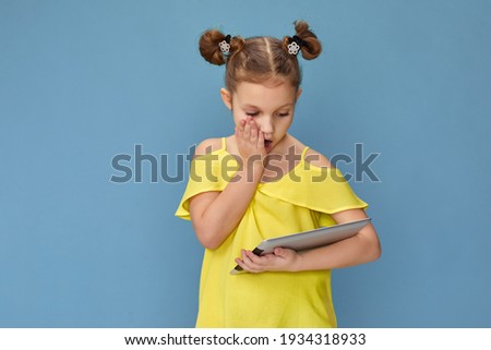 Surprised caucasian child girl in yellow t-shirt using digital tablet on blue background