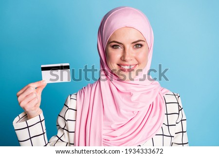 Close-up portrait of pretty content cheerful muslimah girl holding in hand bank card finance isolated over bright blue color background