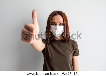 Beautiful caucasian young woman with disposable face mask. Protection versus viruses and infection. Studio portrait, concept with white background. Woman showing thumb up.