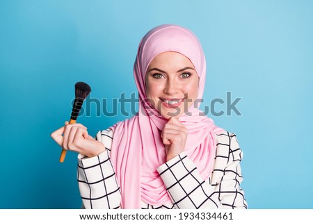 Close-up portrait of lovely cheerful muslimah girl holding in hand brush decorative rouge isolated over shine blue color background