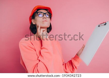 Pretty thoughtful  woman worker builder hold white sign board blank against pink background. Building helmet. 