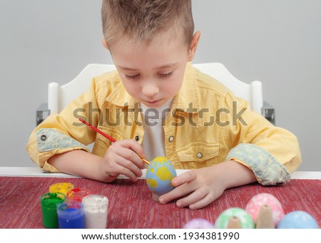 Preschooler boy focused on decorating the Easter egg with a paintbrush and colored paints while sitting at the table at home. Easter family holiday celebration at home and craft concept.