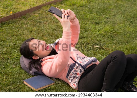 young happy Asian woman in city park - outdoors lifestyle portrait of  cheerful and pretty Korean girl taking selfie with mobile phone relaxed and playful on green grass 
