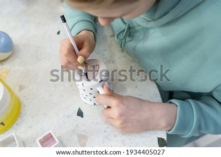 A young woman in casual clothes paint a pink heart on the back of a ceramic cup in a ceramic workshop. Learning new skills, improving skills, hobbies. Ceramic art concept