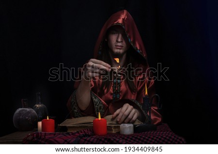Photo poster. The concept of the office of a medieval alchemist: a young magician in a red mantle with a hood sits at a table, in front of him an old tome, burning candles and dusty glass vessels.