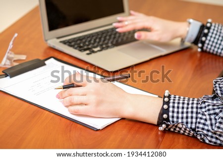 Female hands holding pen under the document and working with laptop at the table in an office