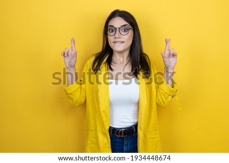 Young brunette businesswoman wearing yellow blazer over yellow background gesturing finger crossed smiling with hope and looking side