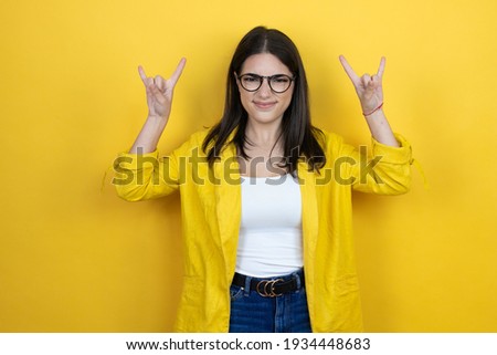 Young brunette businesswoman wearing yellow blazer over yellow background shouting with crazy expression doing rock symbol with hands up