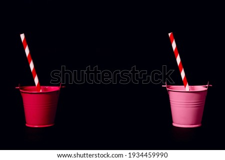 Two buckets on a black background with striped red and white straws. A red and pink bucket around the edges of the photo with free space for text in the center. Cool party or thirsty hot day concept