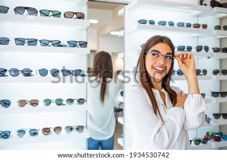 Happy female optometrist, optician is standing with set of glasses in background in optical shop. Stand with spectacles. Girl wearing glasses. Portrait of a woman in correcting glasses. Royalty-Free Stock Photo #1934530742