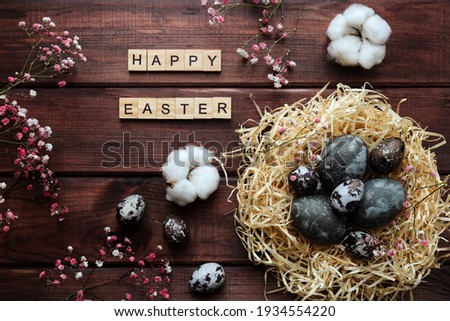Easter greeting card. Nest with naturally dyed eggs, cotton flowers decor and and inscription Happy Easter made from wooden letters on dark wooden background with copy space. Flat lay, top view.