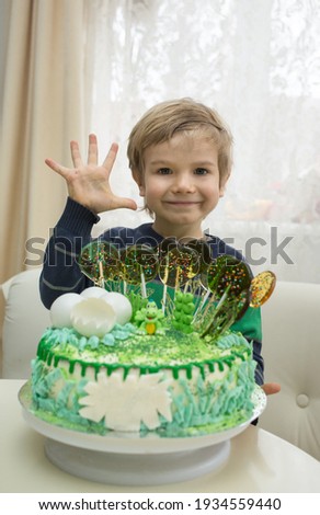 five-year-old boy on his birthday with a beautiful festive cake in green tones. Happy cute little kid smiling, looking at the camera and showing 5 fingers. Celebrating birthday at home.