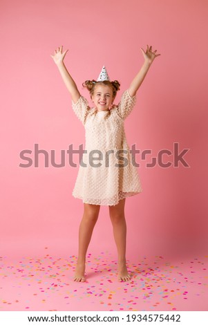 Happy little girl in a birthday cap blows off her palms multicolored confetti on a pink background in the studio