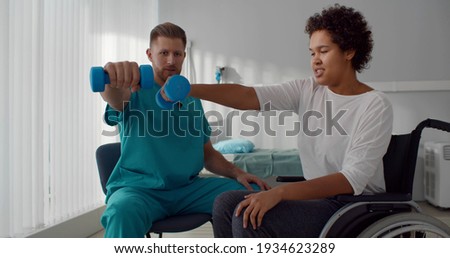 Doctor helping african woman in wheelchair outstretching arms with dumbbells during rehabilitation exercise in modern medical center. Disabled afro-american female having physiotherapy, Royalty-Free Stock Photo #1934623289