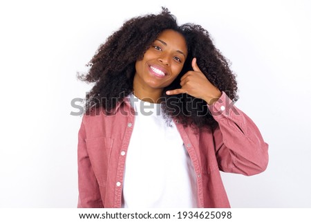 young beautiful African American woman wearing pink jacket against white wall smiling doing phone gesture with hand and fingers like talking on the telephone. Communicating concepts.