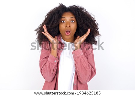 Surprised terrified young beautiful African American woman wearing pink jacket against white wall Gestures with uncertainty, stares at camera, puzzled as doesn't know answer on tricky question, People