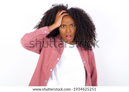 Embarrassed young beautiful African American woman wearing pink jacket against white wall with shocked expression, expresses great amazement, Puzzled model poses indoor