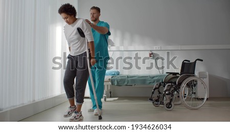 Male medical nurse assisting african young woman walking with crutches. Doctor helping injured woman patient using crutches during rehabilitation in hospital Royalty-Free Stock Photo #1934626034