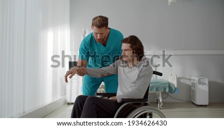 Nurse lifting arm of young paralyzed man sitting in wheelchair. Medical worker stretching arm and doing exercises with disabled male patient in rehabilitation center Royalty-Free Stock Photo #1934626430