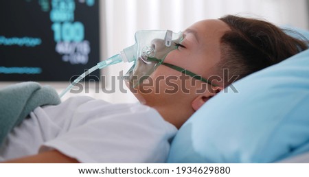 Close up sick young african boy with face oxygen mask sleeping in bed at hospital. Unconscious afro-american kid patient lying in hospital bed with oxygen mask attached to monitor