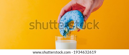 Man hand holding a vibrant and delicious bitten blue doughnut with condensed milk. Dipping into glass of milk. Bright yellow website banner, text copy