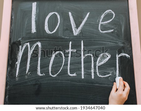 A girl, a child beautifully and diligently writes on a wooden black board, an easel, holding white chalk in her hand, the words and declarations of love for her mother. Photo, copy space.