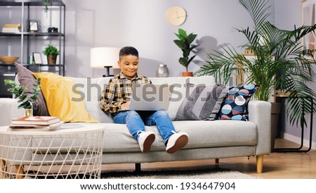 Cheerful cute little African American boy child sitting on sofa in cozy living room at home and browsing online on laptop watching cartoons and smiling, happy childhood, kid concept,