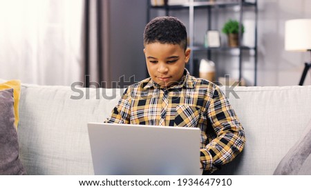 Portrait of cute little African American boy kid sitting on sofa in living room and browsing online on laptop watching cartoons or doing homework, childhood concept, child typing on computer at home
