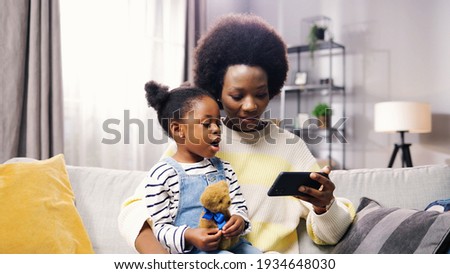Portrait of happy African American family pretty mother and small adorable daughter sitting on sofa browsing online on smartphone watching cartoons. Mom and child searching internet on cellphone