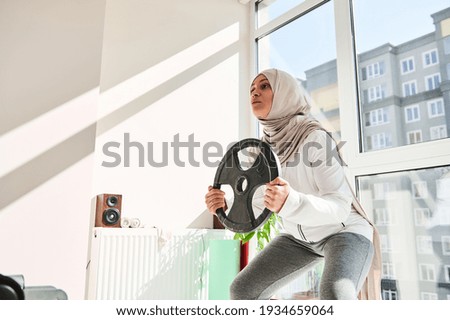 Arab muslim woman with headdress wearing hijab doing weighted squats with a metal disc in her hands - home workout concept.