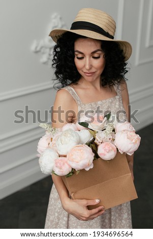 Pink and white peonies in a craft box in the hands of a curly brunette in a straw hat