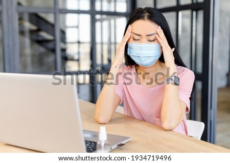 Disappointed female worker holding her head, feeling bad, in medical mask, sitting in the office at the laptop with antiseptic, crisis in the company, keeps distance, lost clients due to the pandemic
