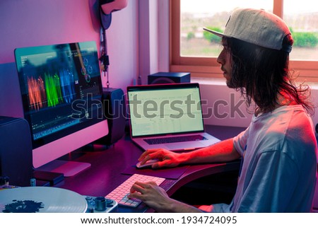 On line video blogger working with a video edition software and looking the stats of his channel in a laptop, with long hair and cap