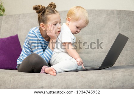 Little cute boy and girl using laptop together, looking at screen, watching cartoons or playing online, sister and brother, siblings sitting on comfortable couch at home, children and gadget concept